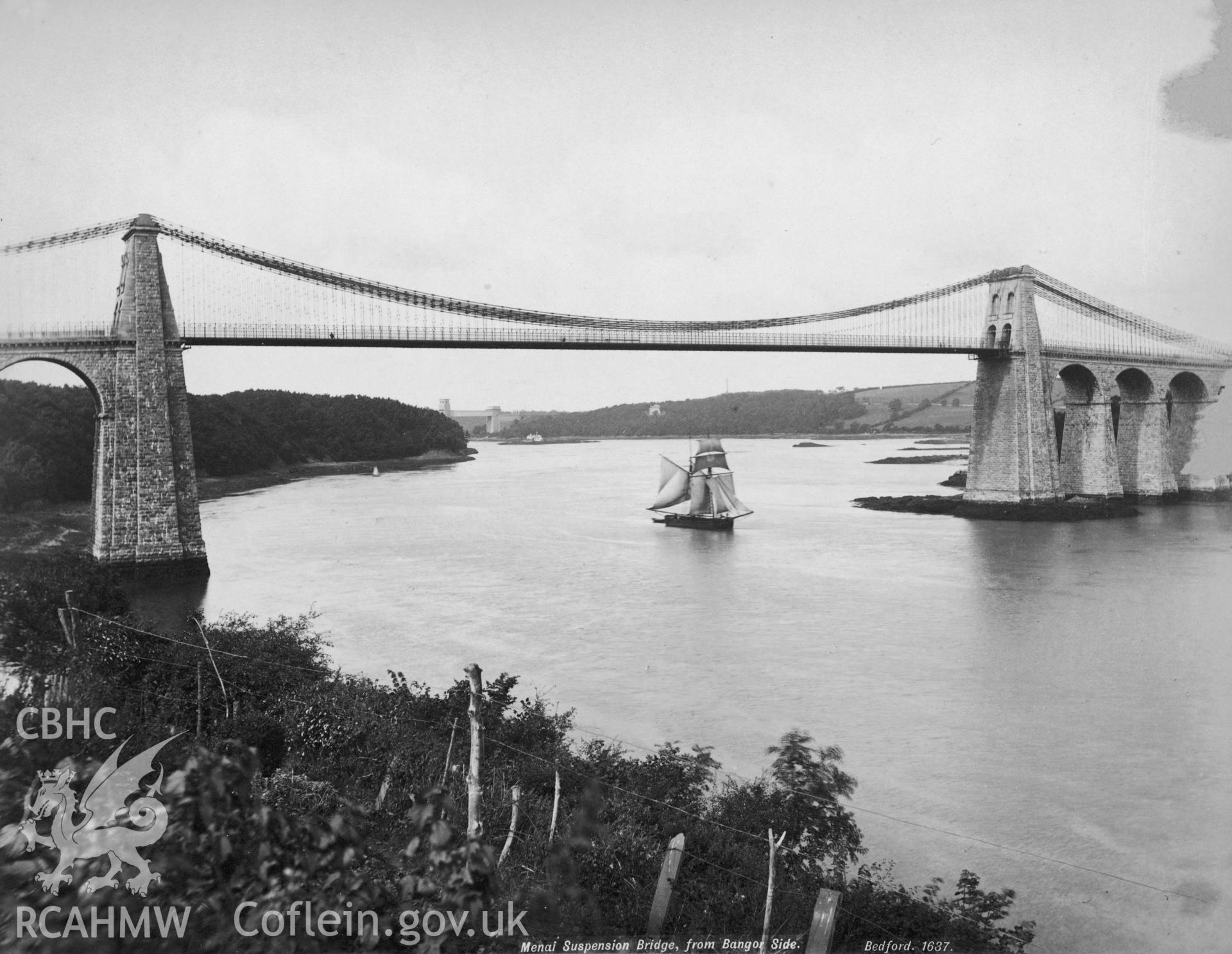 Black and white print of Menai Suspension Bridge, Menai Bridge, showing the bridge from the Bangor side with a topsail schooner sailing underneath.