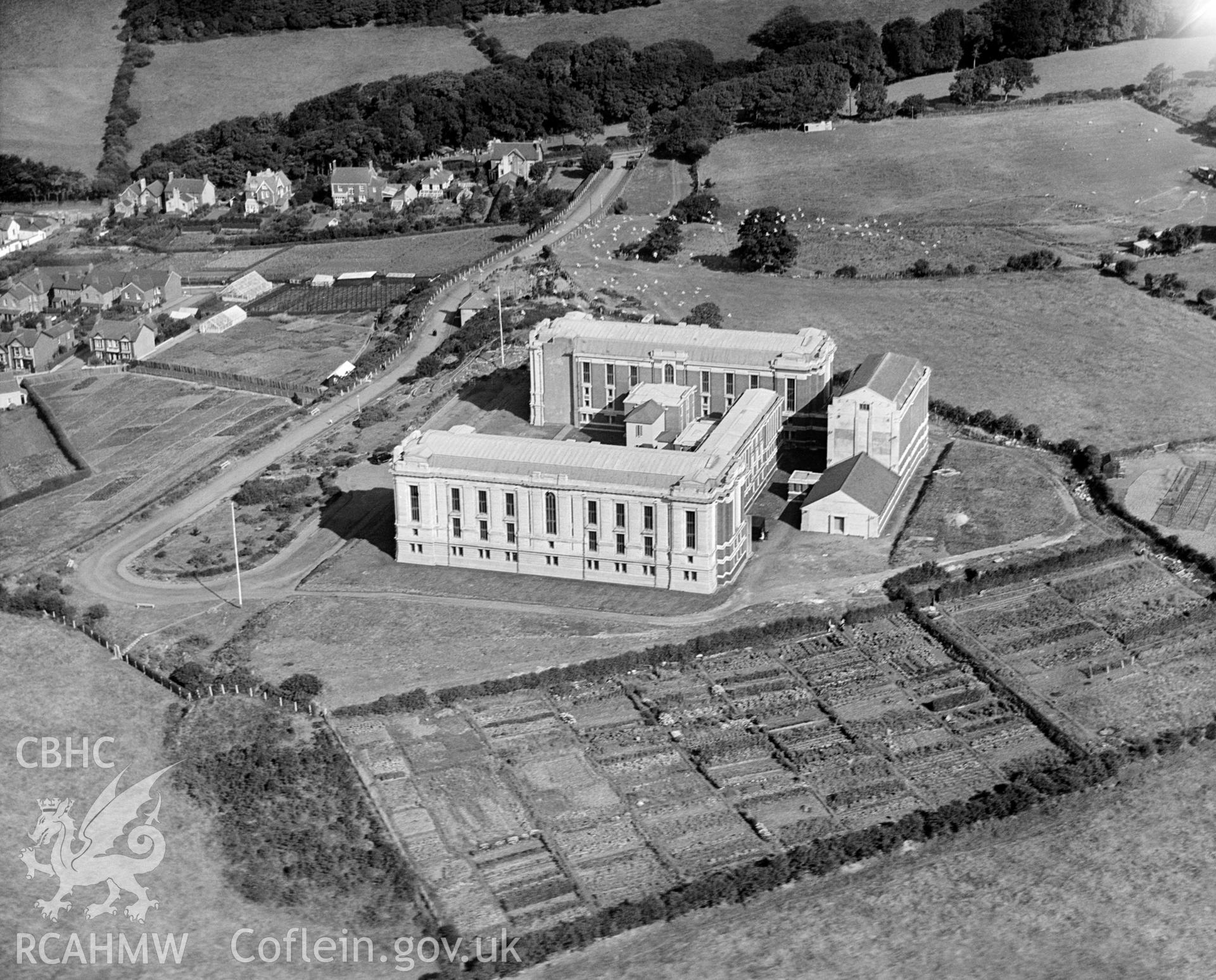 Digital copy of a black and white, oblique aerial photograph of the National Library of Wales, Aberystwyth. The photograph shows the view from the South East, before the main West facade was built and also showing the library's allotment gardens, now covered by a car park.